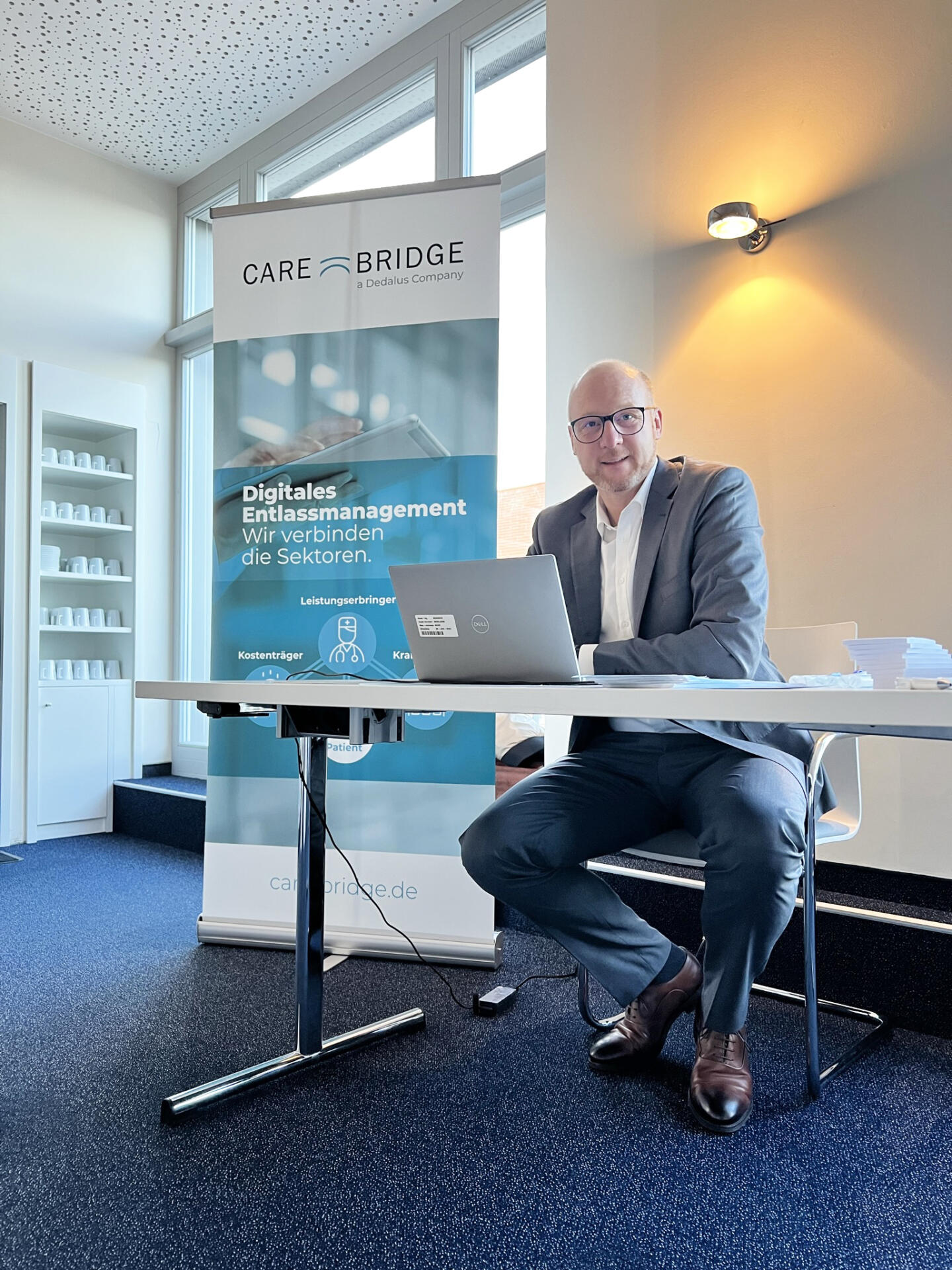 Lars Fehmer sitzt vor seinem Laptop an einem Schreibtisch. Hintergrund ist das Fachsymposium Entlassmanagement in Leipzig.