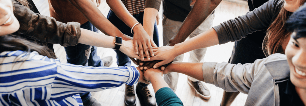 Eine Gruppe von Menschen stehen im Kreis und strecken ihre Hände in die Mitte. Das Bild suggeriert Zusammenhalt und Teamarbeit.