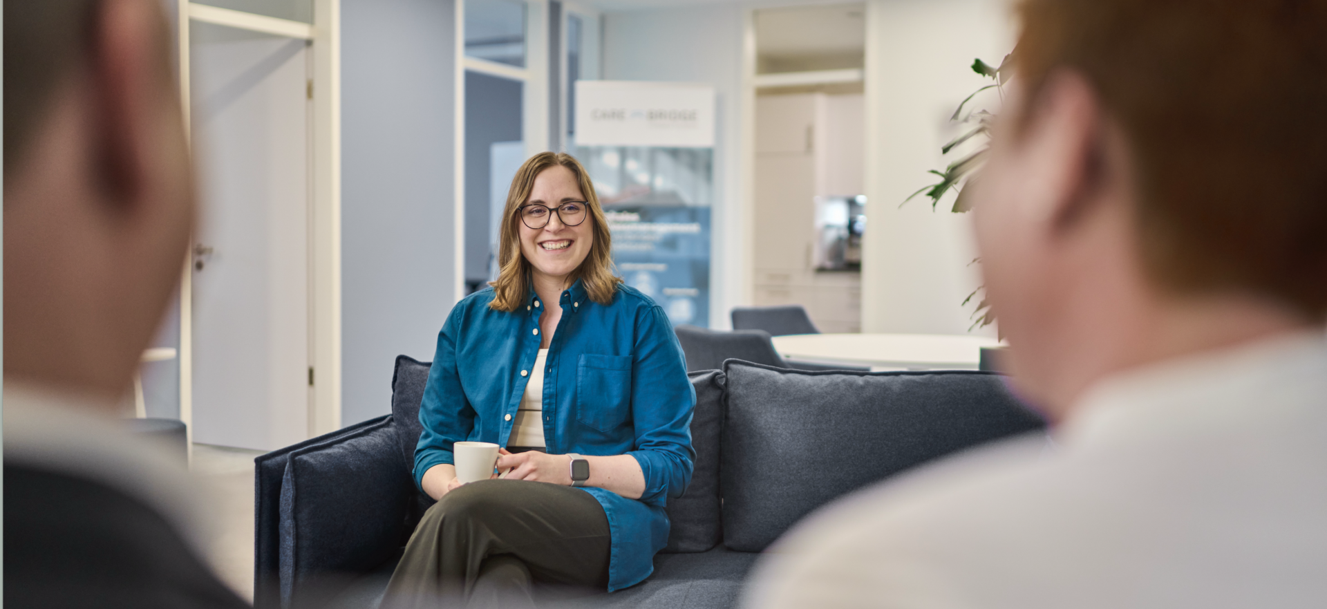 Drei Personen verbringen ihre Mittagspause miteinander. Sie sitzen auf einer Couch im Büro und trinken gemeinsam Kaffee.